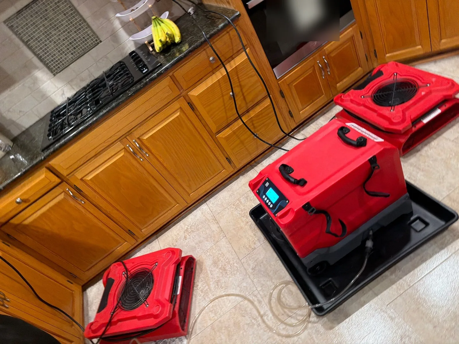 Dehumidifier in a drip pan and air movers set up on a kitchen tile floor to dry a water leak