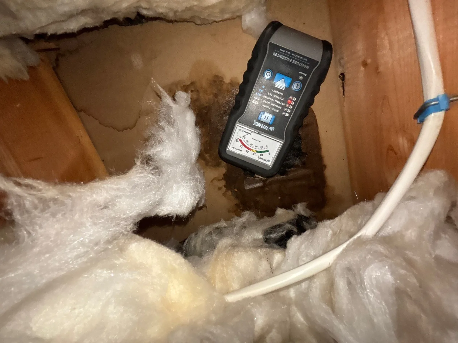 Moisture meter held against a stained drywall cavity above wet, matted fiberglass insulation