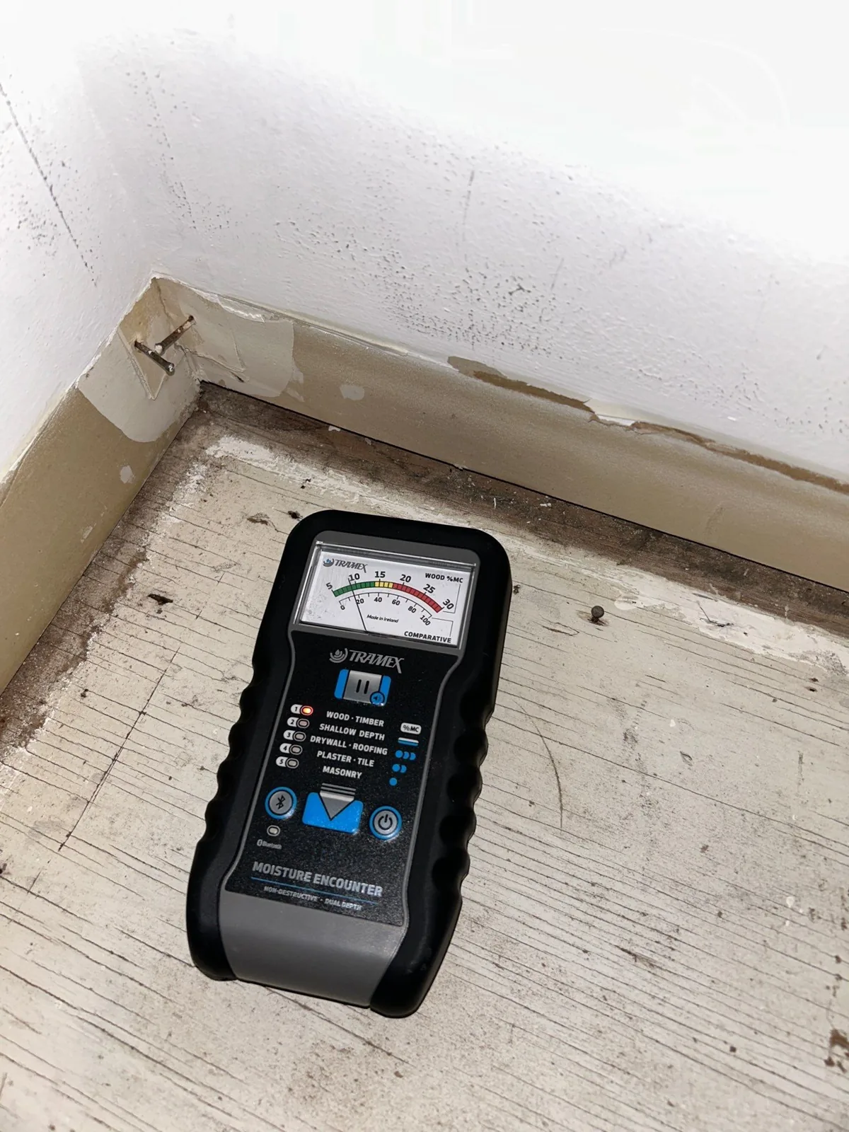 Pinless moisture meter resting on a stripped wood subfloor next to a wall base during a drying check