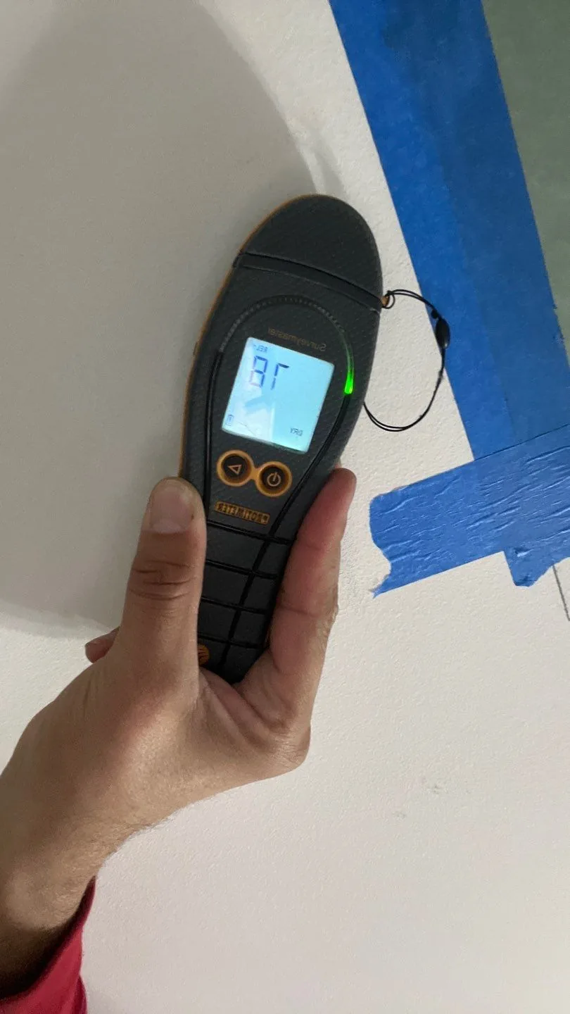 Hand-held moisture meter showing a reading on a wall marked with blue tape