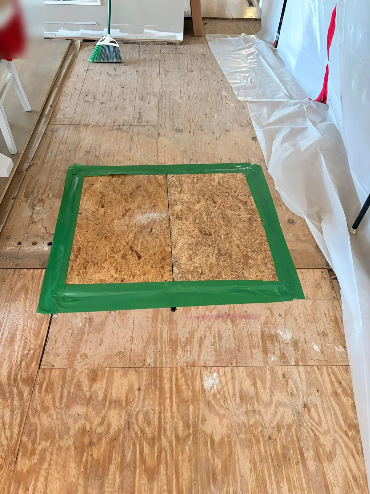 New OSB subfloor panel taped off in green tape within a plywood kitchen subfloor, with plastic sheeting protecting the walls