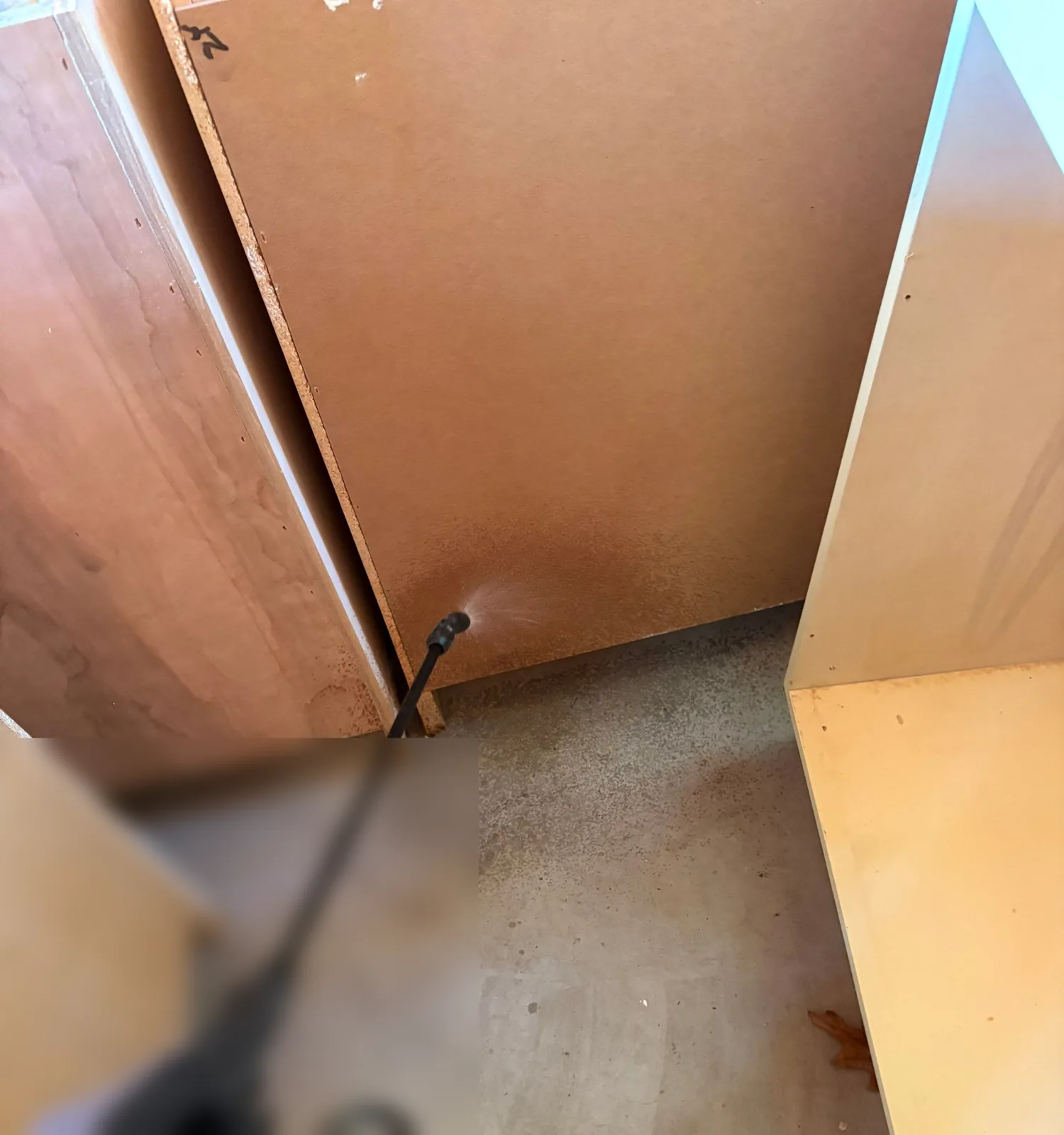Pump sprayer wand applying antimicrobial to the base of a removed-toe-kick kitchen cabinet over a bare concrete floor